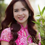 A smiling woman in a pink floral dress poses outdoors surrounded by greenery.